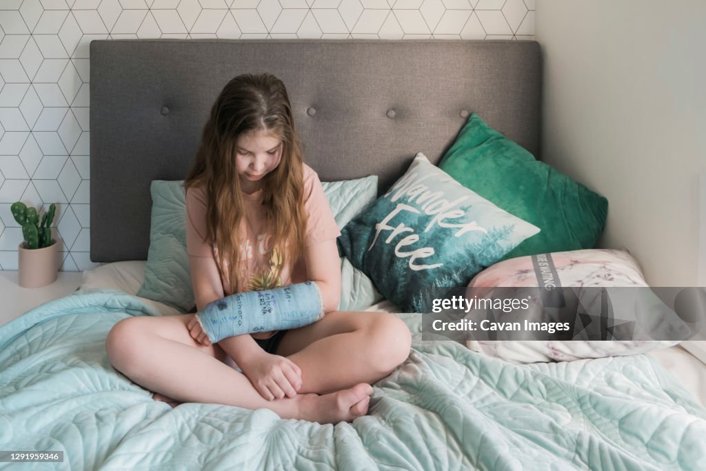 Young girl sitting on a bed looking at her arm in a blue plaster cast