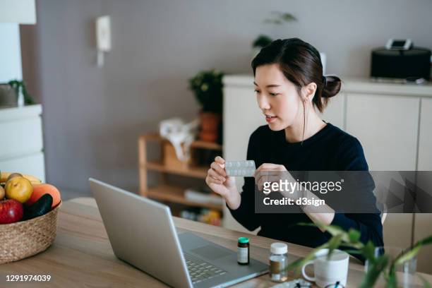 young asian woman video conferencing with laptop to connect with her family doctor, consulting about medicine during self isolation at home in covid-19 health crisis - zorgverzekering stockfoto's en -beelden