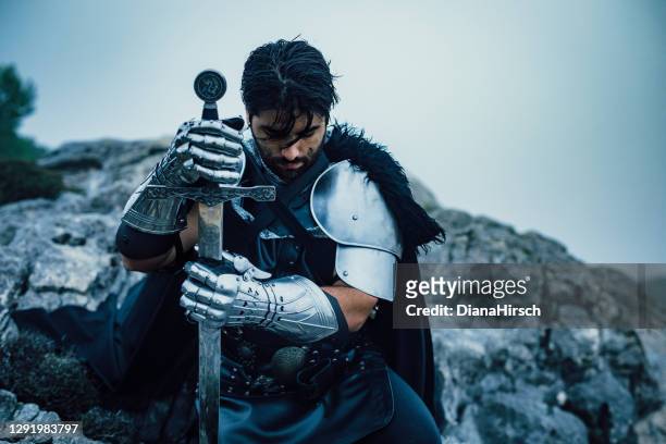 deep feeling portrait of genuflecting praying knight in the mountains after the battle - holding sword stock pictures, royalty-free photos & images