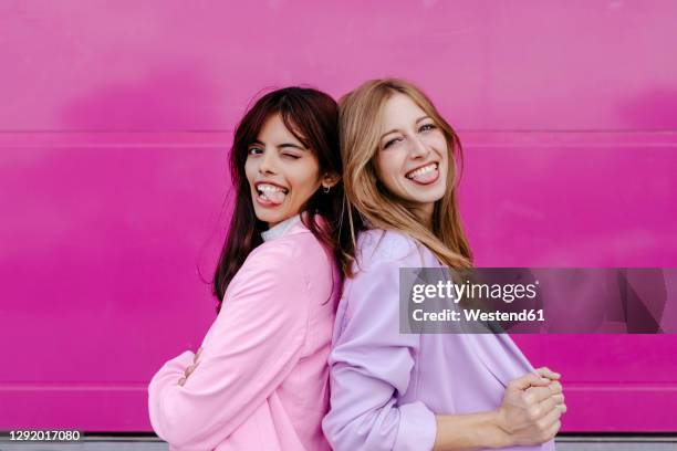 sisters sticking out tongue while standing back to back by pink wall - blond haar stock-fotos und bilder