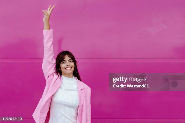 young woman with hand raised doing peace sign while standing against pink wall - 手を挙げる ストックフォトと画像