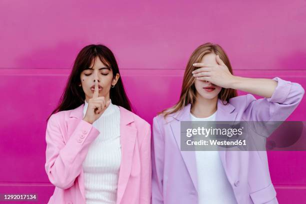 young woman with finger on lips standing by sister covering eyes against pink wall - be silent stock-fotos und bilder