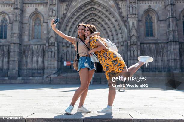 woman embracing friend while taking selfie through mobile phone standing at barcelona cathedral square in barcelona, catalonia, spain - barcelona cathedral stock pictures, royalty-free photos & images