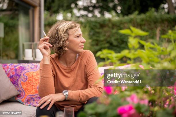 woman holding cigarette while sitting on sofa at backyard - femme et fumeuse photos et images de collection