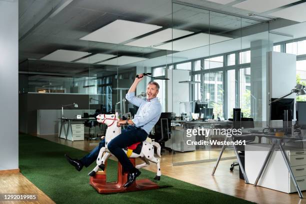 cheerful businessman looking away while sitting on horse toy at office - schaukelpferd stock-fotos und bilder