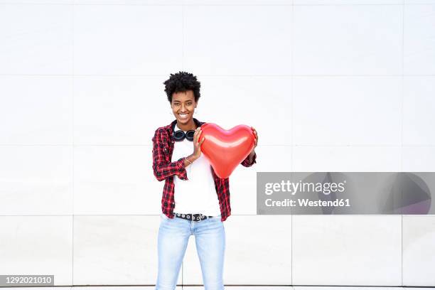 happy young female hipster holding red heart shape balloon against white wall - black balloon stock-fotos und bilder