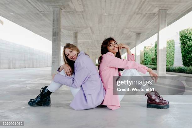 happy siblings sitting back to back on floor in parking lot - lila blazer stock-fotos und bilder