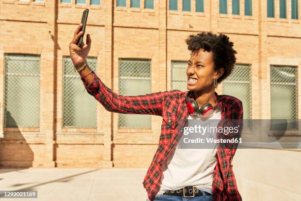 young woman clenching teeth while taking selfie through mobile phone against building - open hair selfie stock pictures, royalty-free photos & images
