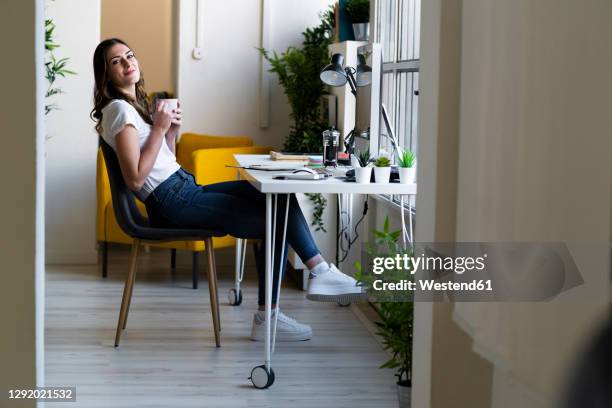 smiling businesswoman holding coffee cup while sitting on chair at office - table side view stock pictures, royalty-free photos & images