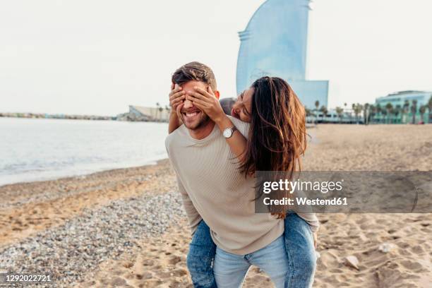 boyfriend giving piggyback ride while girlfriend covering his eyes at beach - augen zuhalten stock-fotos und bilder