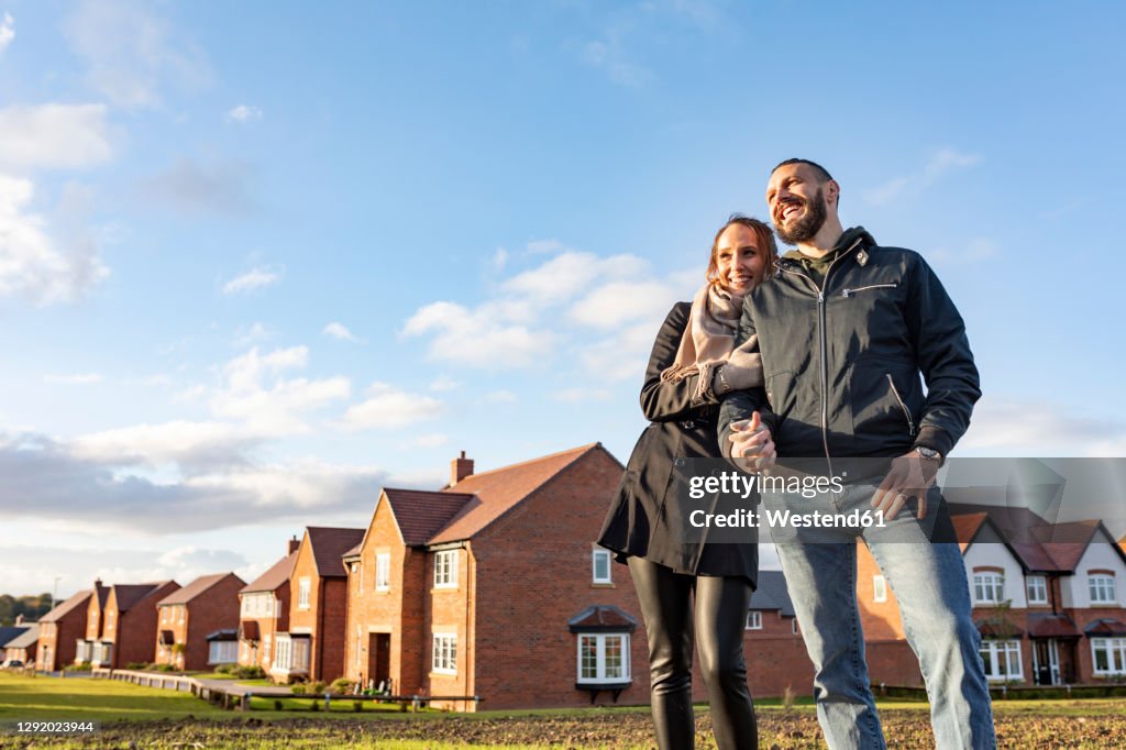 Happy couple looking away while standing outside newly built houses against sky