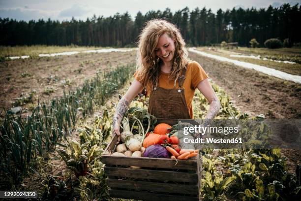 smiling farm worker carrying vegetable box while standing at farm - farm worker foto e immagini stock