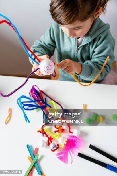 cute girl making toys of styrofoam ball and pipe cleaners while sitting at table in living room - lesseps stockfoto's en -beelden