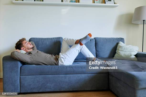 man resting on sofa with hands behind head in living room at home - überkreuzte beine stock-fotos und bilder