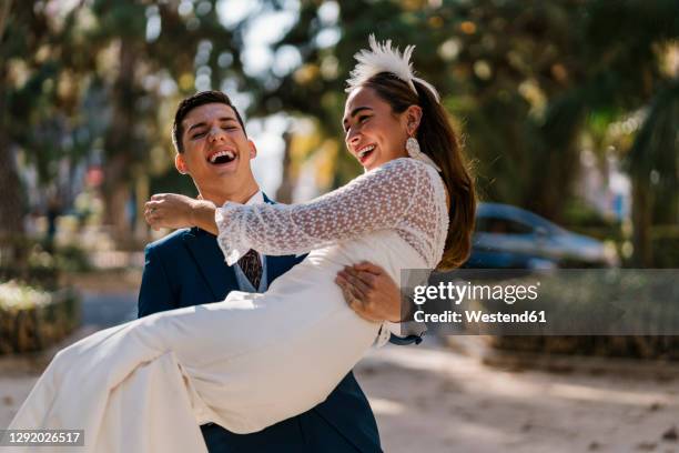 bride laughing while carrying groom in park on sunny day - bride and groom stock-fotos und bilder