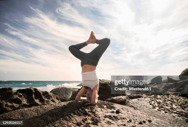 woman practicing sirsasana on rock formation at beach against sky - shirshasana stock pictures, royalty-free photos & images