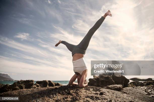 carefree woman practicing sirsasana with legs apart on rock formation at beach against sky - shirshasana stock pictures, royalty-free photos & images
