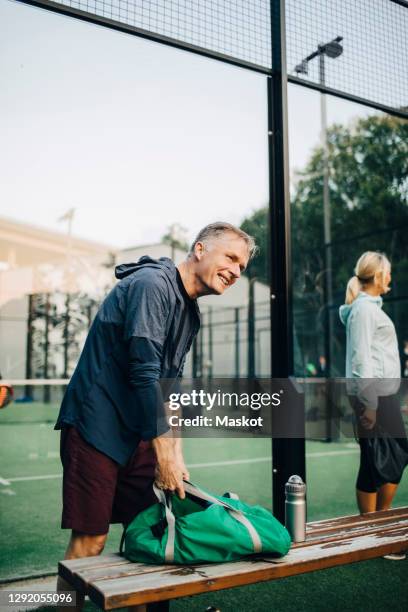 smiling senior man with bag standing by bench in sports court - banco de jogadores fotografías e imágenes de stock