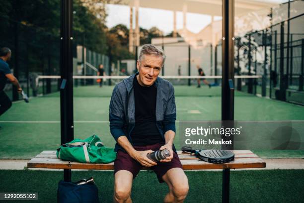 portrait of smiling retired man with bottle sitting on bench in sports court - match sport stock-fotos und bilder