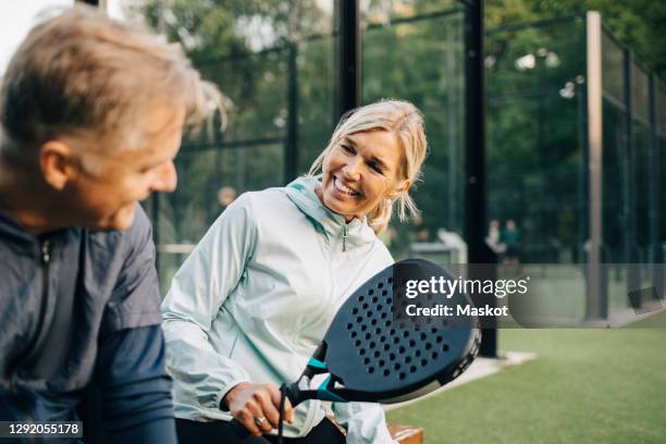 smiling female with racket talking to friend while sitting in sports court - paddle tennis stock pictures, royalty-free photos & images