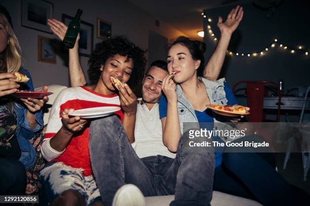 friends eating pizza on sofa during party at home - vriendin stockfoto's en -beelden