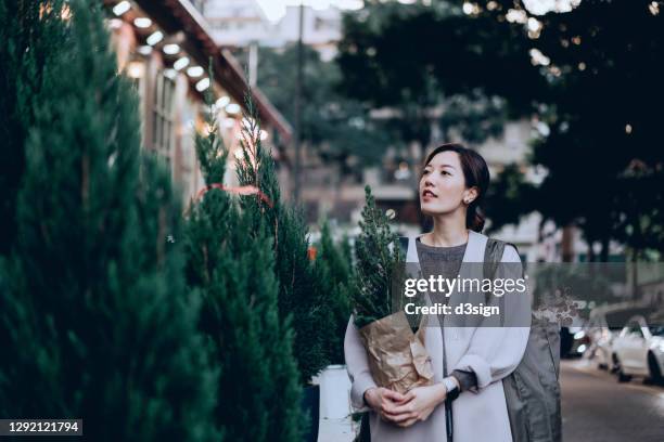 young asian woman shopping in the flower market in the city, carrying a pine tree and a eucalyptus with a reusable shopping bag. preparing for christmas. christmas holiday vibe. christmas lifestyle theme - sustainability asia stock pictures, royalty-free photos & images