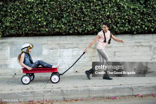 cheerful girl pulling friend sitting in wagon on sidewalk - helmet cart stock pictures, royalty-free photos & images