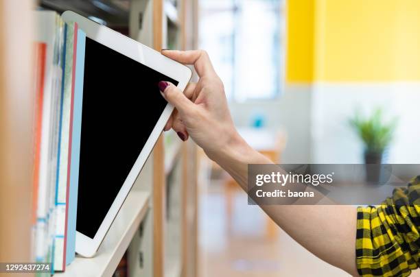 woman picking digital tablet from bookshelf - digital handhold device stock pictures, royalty-free photos & images