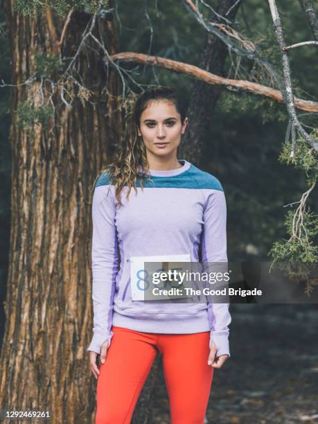 portrait of confident female athlete with marathon bib standing by tree in forest - startnummer stock-fotos und bilder