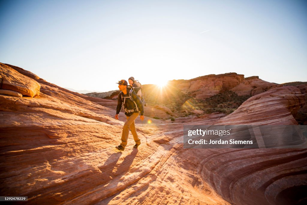 A man carrying his son in a backpack while hiking in the red rocky desert landscape.