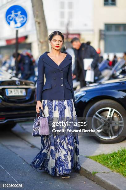 Thassia Naves wears earrings, a navy blue double-breasted jacket with a large cleavage, a long flowing white and blue floral print pleated skirt, a...
