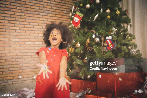 african american child excitedly throwing glitter and playing with christmas decoration gathered near christmas tree - sparkle children stock pictures, royalty-free photos & images
