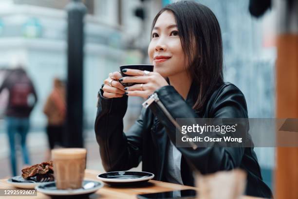 confident and smiling young asian woman enjoying coffee at sidewalk cafe - アフタヌーンティー ストックフォトと画像