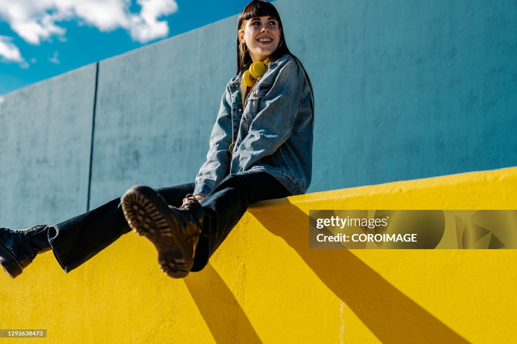 Smiling young woman sitting on a yellow wall in sunny day