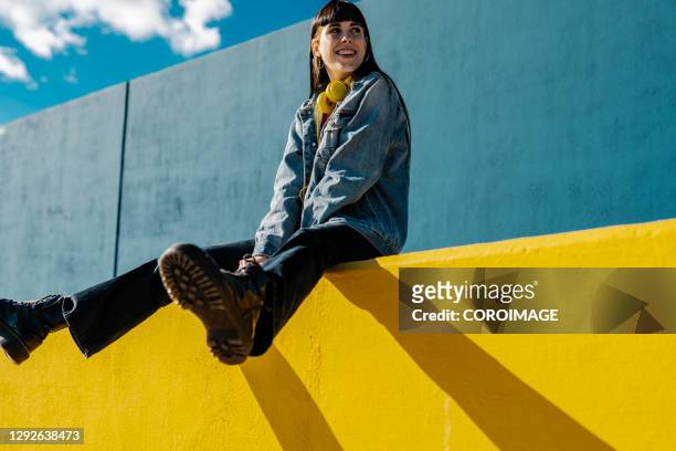 smiling young woman sitting on a yellow wall in sunny day - städtische straße stock-fotos und bilder