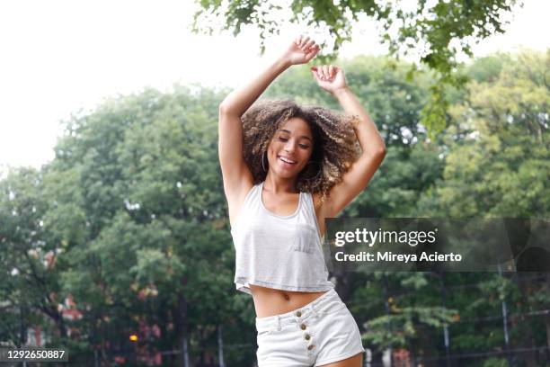 a beautiful latinx millennial woman with curly light brown hair, happily dances in the park wearing a light grey tank top and jean shorts. - woman vest stock pictures, royalty-free photos & images