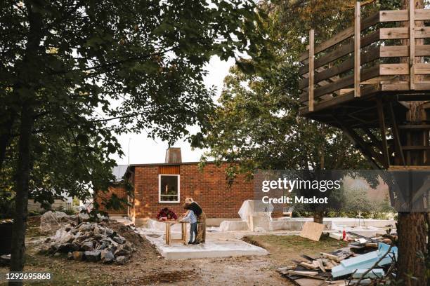 mother and son using hand saw while father with daughter by concrete wall against house during summer - backyard renovation stock pictures, royalty-free photos & images