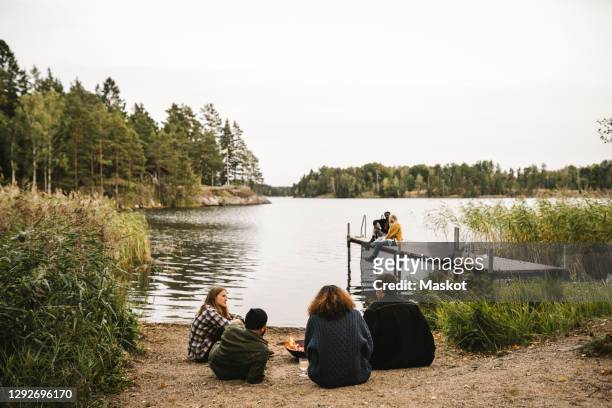 rear view of male and female talking friends while couple sitting on pier during social gathering - stockholm beach stock pictures, royalty-free photos & images