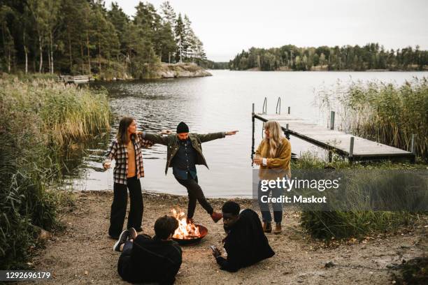 male and female dancing by fire pit while friends sitting near lake during social gathering - male friends hanging out stock-fotos und bilder