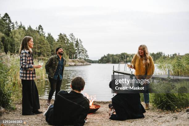 male and female friends enjoying by fire pit near lake during social gathering - stockholm beach stock pictures, royalty-free photos & images