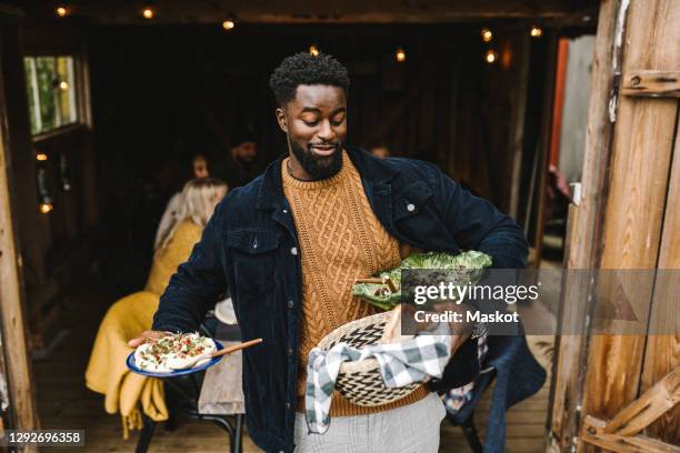 smiling man balancing food while standing at doorway during social gathering - reunião de amigos imagens e fotografias de stock