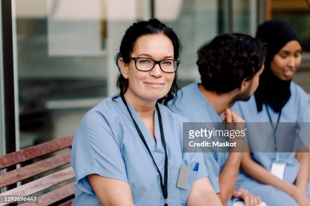 portrait of smiling female nurse in eyeglasses sitting by colleagues on bench - nurse headshot stock pictures, royalty-free photos & images