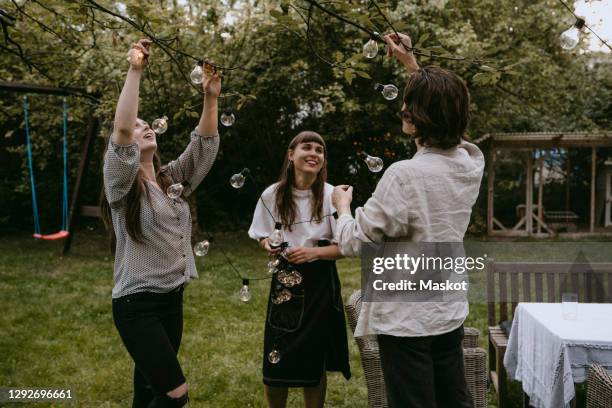 smiling friends hanging lighting equipment in yard during dinner party - hanging in garden photos et images de collection