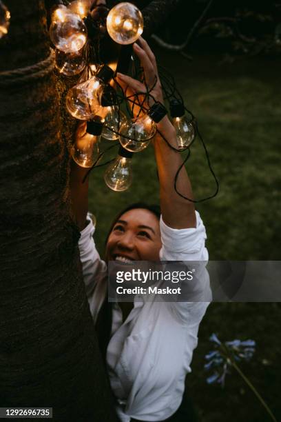 smiling female hanging lighting equipment on tree trunk in yard during dinner party - summer lights stockfoto's en -beelden