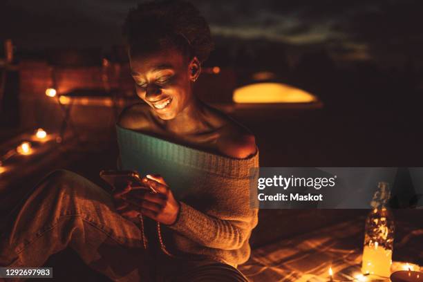 happy woman using mobile phone while sitting on picnic blanket at rooftop - rooftop party night stock pictures, royalty-free photos & images