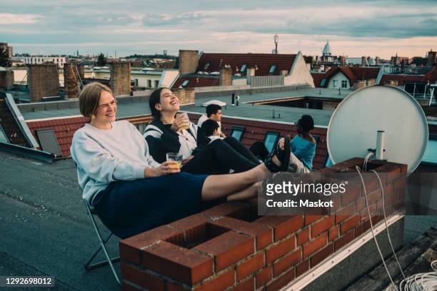 smiling female talking to friend while enjoying drink on building terrace - young adult stock-fotos und bilder