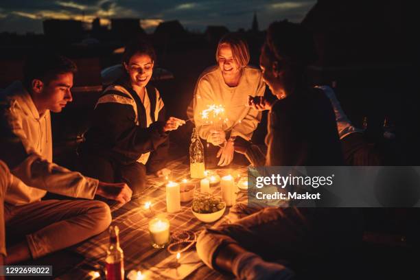 smiling female and male friends igniting sprinkler on building terrace at night - night picnic stock pictures, royalty-free photos & images