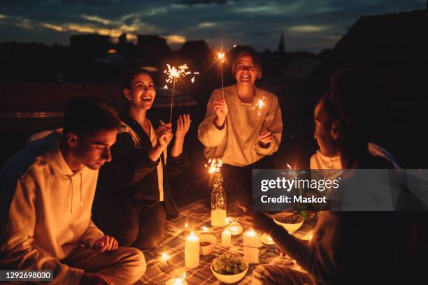 smiling female and male friends igniting sprinkler on building terrace at night - night picnic stock-fotos und bilder