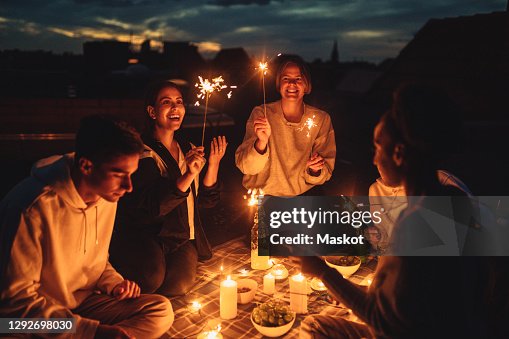 Smiling female and male friends igniting sprinkler on building terrace at night Smiling female and male friends igniting sprinkler on building terrace at night