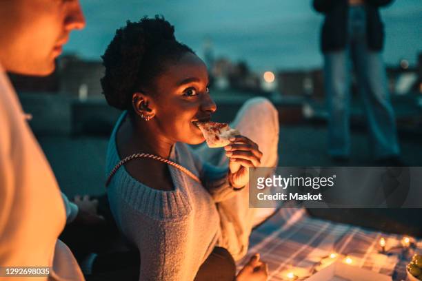smiling female eating pizza by friend on rooftop at night - adult photo sharing stock pictures, royalty-free photos & images